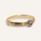  Gold cuff bracelet with ridged band and clear round gemstone at center.